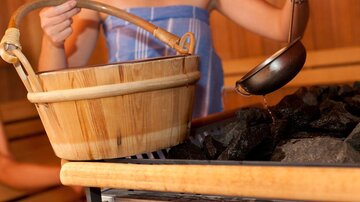 A person is pouring water from a ladle onto hot stones in a sauna. Next to it stands a wooden basket.