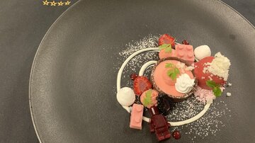 An artistically arranged dessert on a black plate. It consists of various sweet components with a pink cream, fruits, and a decorative whipped cream.