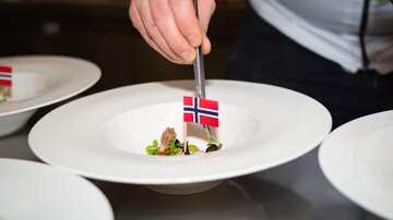 A chef decorates a plate with a small Norwegian flag symbol. The dish consists of meat and fresh herbs.
