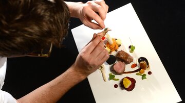 A person artfully arranges a gourmet dish on a white plate. The presentation includes various ingredients and sauces in vibrant colors.