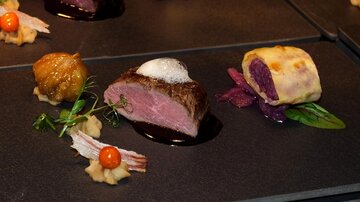 An artfully arranged plate with grilled meat, side dishes, and sauces. The presentation showcases various textures and colors on a dark background.