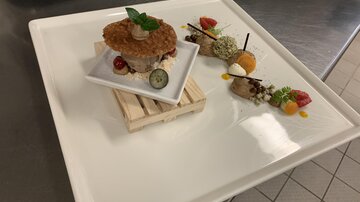 An artistically arranged plate with a main dish in a bowl on a small wooden board. Next to it, various creative sides and sauces are arranged.