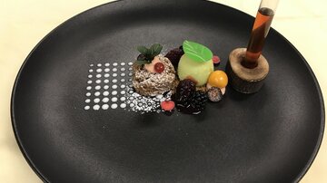 An artistically arranged dessert on a black plate. It contains various fruits, a green cream, and a small glass tube with a liquid.