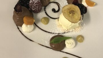 An artistic dessert on a plate, decorated with chocolate sauce and various fruits. It contains chocolate mousse, whipped cream, and banana slices.