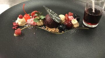 An artistically arranged dessert on a black plate. It consists of various fruits, creams, and a small portion of drink.