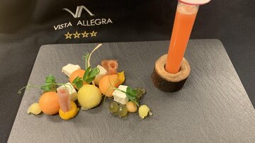 An artistically arranged dish on a black plate, consisting of various colorful ingredients and a small drink column. It features a mix of melons, cheese, and decorative surface elements.