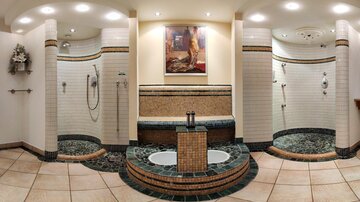 An elegant bathroom with multiple shower areas and a decorative sink. The walls are designed with ceramic tiles and there is an artistic wall decoration.