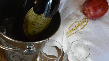 A bottle of champagne is standing in an ice bucket, accompanied by two glasses and a red apple. In the background, there is a towel with a logo.