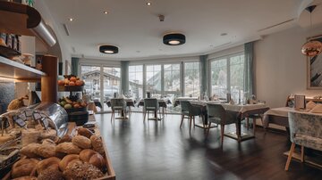 A modern restaurant with wooden flooring and elegant tables. In the foreground, fresh rolls and fruit can be seen, while large windows offer a view of the mountains.