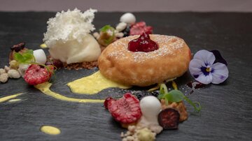 An artfully arranged dessert on a black plate. It consists of a baked pastry, fresh berries, a scoop of ice cream, and edible flowers.