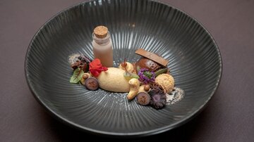 A beautifully arranged plate with various desserts. The dish includes a bottle with a cream, different fruits, and chocolate elements.