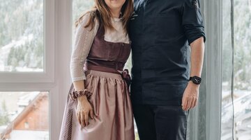 A couple stands in a room with large windows that display a mountain landscape. The woman is wearing a dirndl, while the man is dressed in a dark shirt.