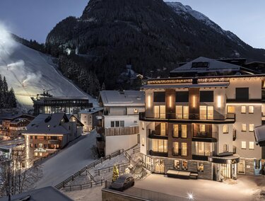 Ein Winterszene mit einem beleuchteten Hotel in den Bergen. Im Hintergrund sind schneebedeckte Pisten und ein hübsches Dorf zu sehen.