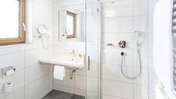 Ein modernes Badezimmer mit einer gläsernen Dusche und hellen Fliesen. Es gibt ein Fenster und eine Waschbeckenablage.