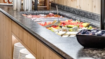 Ein einladendes Buffet mit frischem Gemüse, Obst und Aufschnitt. Der Bereich ist in einem holzernen, modernen Stil gestaltet.
