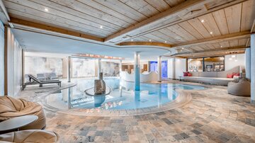 Ein modernes Schwimmbad in einem stylischen Innenraum mit Holzdecke. Bequeme Sitzgelegenheiten und ein entspannender Bereich laden zum Verweilen ein.