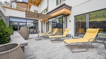 Eine moderne Terrasse mit gelben Liegen und einem Sonnenschirm. Im Hintergrund sind große Fenster und ein elegantes Wohngebäude zu sehen.