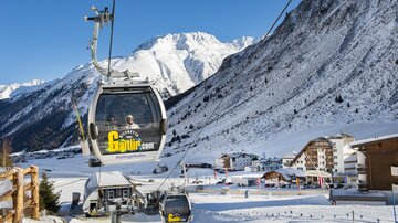 Eine Gondelbahn fährt durch eine schneebedeckte Landschaft mit hohen Bergen. Im Vordergrund sind Gebäude und Skipisten sichtbar.