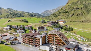 Eine malerische Berglandschaft mit mehreren Gebäuden und grünen Wiesen. Die Sonne scheint und der Himmel ist klar.