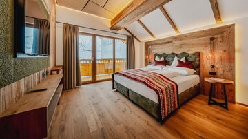 Ein modernes Schlafzimmer mit Holzdekor und großen Fenstern. Das gemütliche Bett ist mit roten und grünen Kissen dekoriert und bietet einen Blick auf die Berge.