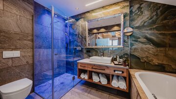 Ein modernes Badezimmer mit einer gläsernen Dusche und einer eleganten Badewanne. Der Holzschrank mit Handtüchern und der bunte Stein auf der Wand verleihen einen stilvollen Look.