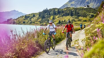 Zwei Radfahrer fahren auf einem schönen Waldweg entlang eines Sees. Die Umgebung ist grün und bergig, mit bunten Blumen am Wegesrand.