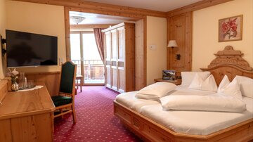 Ein gemütliches Hotelzimmer mit Holzmöbeln und einem großen Bett. Es gibt einen Fernseher, einen Schreibtisch und einen Balkon mit Blick nach draußen.