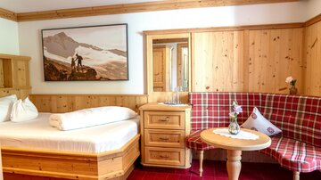 Ein gemütliches Zimmer mit Holzmöbeln und einem aufgeräumten Bett. An der Wand hängt ein Bild von Bergen und einer Person, die die Aussicht genießt.