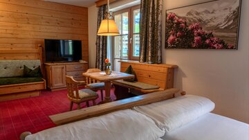Ein gemütlicher Wohnraum mit Holzverkleidung und einem kleinen Essbereich. Am Fenster steht eine Blumenvasen und ein Bild einer Berglandschaft hängt an der Wand.