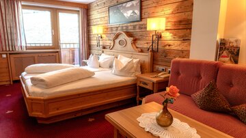 Ein gemütliches Hotelzimmer mit einem großen Doppelbett und einem Sofa. Der Raum ist mit Holzverkleidung und einem Teppich dekoriert und bietet einen Blick auf die Berge.