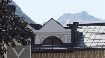 Eine Seilbahn schwebt über einem traditionellen Gasthof in den Bergen. Im Hintergrund sind die Gipfel der Berge sichtbar.