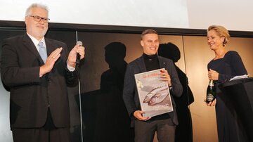 Drei Personen stehen auf einer Bühne. Der Mann in der Mitte hält eine Auszeichnung und wird von den anderen applaudiert.